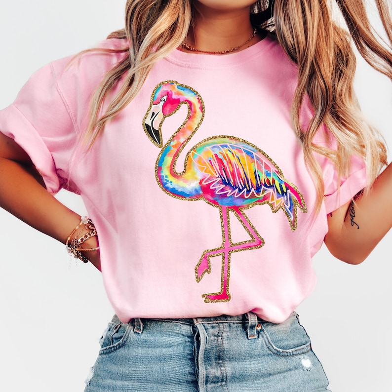 May include: Pink sweatshirt featuring a colorful flamingo design. The flamingo has a rainbow-colored body with gold glitter accents. The bird is standing on one leg. The sweatshirt is a solid pink color.