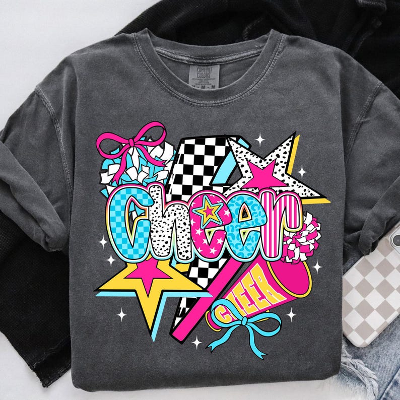 Op de afbeelding: Donkergrijze sweatshirt met een kleurrijke "Cheer"-afbeelding. Het ontwerp bevat een ster, pompons en een megafoon, met het woord "Cheer" in een mix van patronen en kleuren, waaronder roze, blauw en zwart.