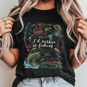 May include: Black t-shirt with a colorful fishing-themed graphic. The design includes fishing rods, fish, a tackle box, and the text "I'd rather be fishing". The illustration features a boat and lily pads.