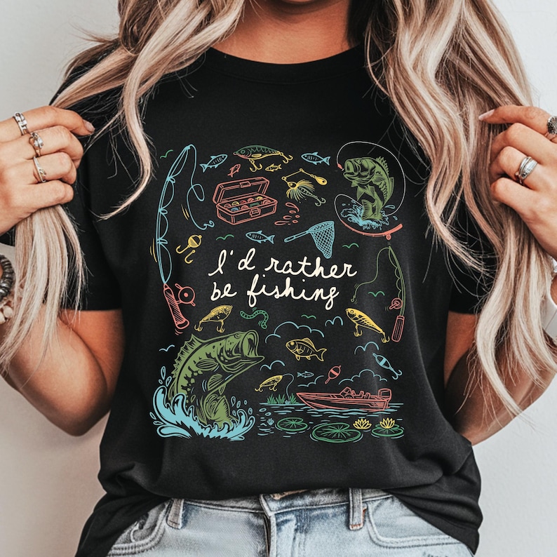 May include: Black t-shirt with a colorful fishing-themed graphic. The design includes fishing rods, fish, a tackle box, and the text "I'd rather be fishing". The illustration features a boat and lily pads.