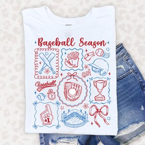 May include: White t-shirt with a baseball-themed graphic. The design includes baseball bats, a glove, a ball, a stadium, and the text "Baseball Season" in red and blue. The shirt is folded on top of denim shorts.