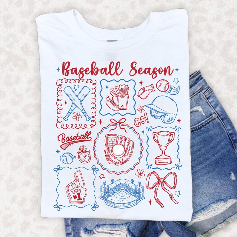 May include: White t-shirt with a baseball-themed graphic. The design includes baseball bats, a glove, a ball, a stadium, and the text "Baseball Season" in red and blue. The shirt is folded on top of denim shorts.