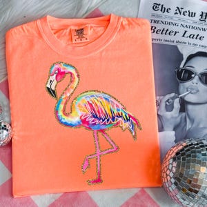 May include: A coral-colored t-shirt featuring a colorful flamingo design with gold glitter accents. The flamingo has a rainbow of colors in its feathers. The shirt has a crew neck and short sleeves. The brand label is visible at the top.