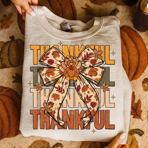 May include: A beige sweatshirt with the word "THANKFUL" in orange, brown, and gray block letters. A large bow with a turkey in the center is the focal point. The bow has a fall leaf pattern in shades of brown, orange, and beige.