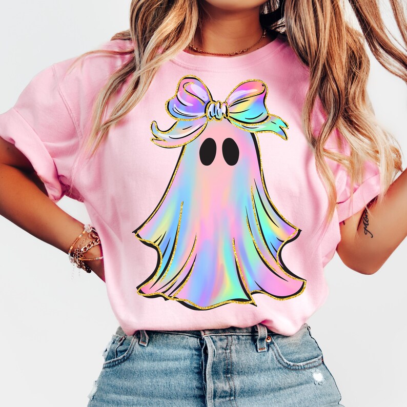 May include: Pink t-shirt featuring a colorful ghost design with a bow. The ghost has a holographic effect with black eyes and gold outlines. The bow is pastel rainbow colored. The shirt is made of a soft material.