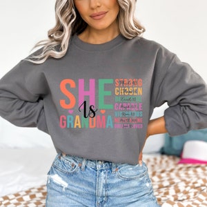 May include: Gray sweatshirt with the text "SHE Is GRANDMA" in rainbow colors. The text is stacked vertically with additional text in smaller font below each letter. The smaller text includes bible verses and dates.