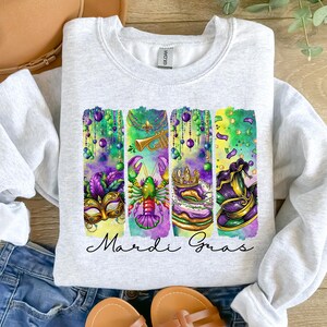 May include: A grey sweatshirt with a colorful Mardi Gras design. The design features a mask, a lobster, a king cake, and a jester's hat. The text "Mardi Gras" is printed below the design.