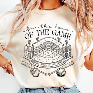 Op de afbeelding: Cr&egrave;mekleurig T-shirt met een zwart-witte honkbalstadionafbeelding en de tekst "for the love OF THE GAME". Het ontwerp bevat honkbalknuppels, een honkbal en een handschoen.