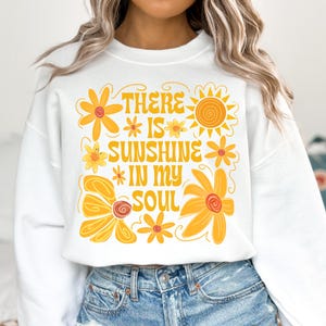 May include: White sweatshirt with a yellow and orange graphic design featuring flowers, a sun, and the text "There is sunshine in my soul."