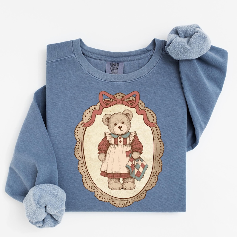 Op de afbeelding: Blauw sweatshirt met een vintage illustratie van een teddybeer die een jurk draagt en een quilt vasthoudt. De beer is omlijst in een ovaal met een rode strik. De sweatshirt heeft een ronde hals en lange mouwen met een zachte, donzige binnenkant.