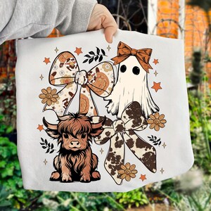 Op de afbeelding: Witte draagtas met een cartoon spook, een Schotse hooglander en twee grote strikken met bloemen- en koeienprintpatronen. Het ontwerp bevat sterren en bloemen in bruin- en oranjetinten.