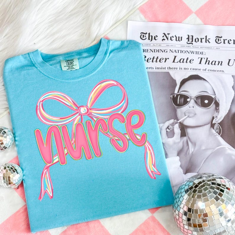 Pode incluir: Uma camiseta azul clara com a palavra "Nurse" em rosa, acentuada por um design de la&ccedil;o colorido. A camiseta &eacute; exibida em uma superf&iacute;cie xadrez rosa e branca, com um recorte de jornal e bolas de discoteca no fundo.