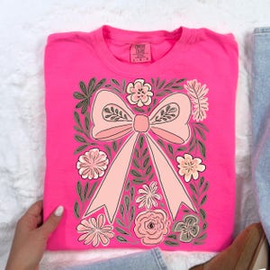 K&ouml;nnte beinhalten: Ein leuchtend pinkfarbenes Sweatshirt mit einem gro&szlig;en, hellrosa Schleifendesign, umgeben von floralen und Blattillustrationen in Rosa-, Gr&uuml;n- und Graut&ouml;nen. Das Sweatshirt hat einen Rundhalsausschnitt und lange &Auml;rmel. Das Comfort Colors-Markenetikett ist sichtbar.