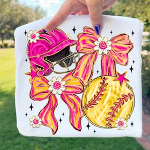 May include: White sweatshirt with a softball theme. Features a pink softball helmet, a yellow softball, and pink and yellow bows. Accented with white flowers and star details. The design is held by a hand.