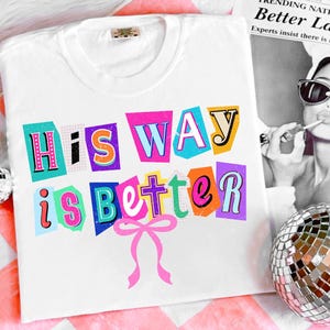 May include: White t-shirt with the phrase "His Way is Better" in colorful, cut-out style letters. A pink bow is below the text. The shirt is displayed on a pink and white striped surface.