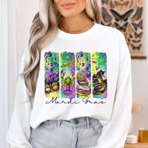 May include: White long-sleeve sweatshirt with a watercolor design of Mardi Gras themed items, including a mask, a crawfish, a king cake, and a hat. The text "Mardi Gras" is printed below the design.