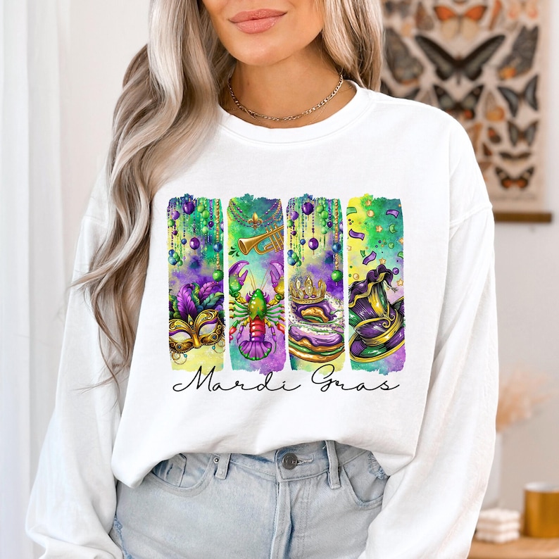 May include: White long-sleeve sweatshirt with a watercolor design of Mardi Gras themed items, including a mask, a crawfish, a king cake, and a hat. The text "Mardi Gras" is printed below the design.