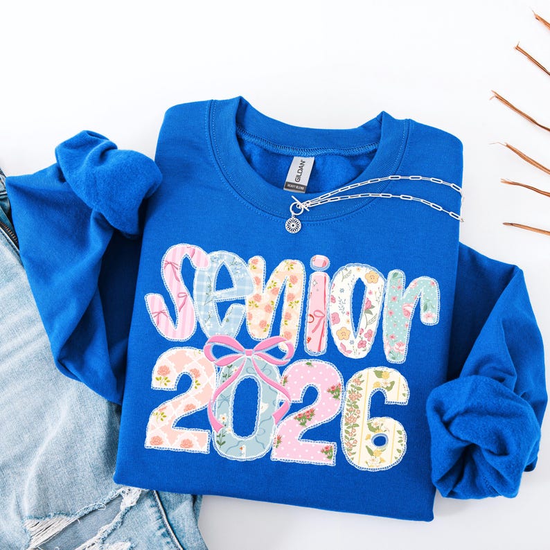 Puede incluir: Sudadera azul real con el texto "Senior 2026" en un dise&ntilde;o de patchwork con patrones florales y pastel. Una cadena de plata con un colgante est&aacute; sobre la camisa. La sudadera est&aacute; doblada sobre unos vaqueros azules.