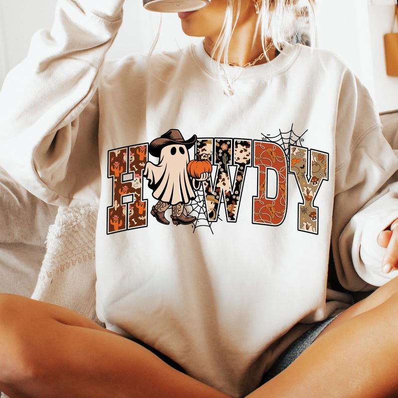 Puede incluir: Sudadera color crema con la palabra "HOWDY" en una fuente de estilo occidental. La "O" se reemplaza con un fantasma con sombrero y botas de vaquero, sosteniendo una calabaza. Tambi&eacute;n hay un gr&aacute;fico de telara&ntilde;a.