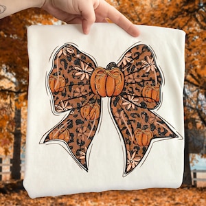 May include: A white sweatshirt featuring a large bow design. The bow has a leopard print pattern with orange pumpkins and floral accents. A large orange pumpkin is in the center of the bow. The design is outlined in black.
