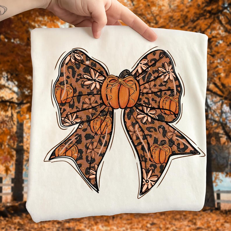 May include: A white sweatshirt featuring a large bow design. The bow has a leopard print pattern with orange pumpkins and floral accents. A large orange pumpkin is in the center of the bow. The design is outlined in black.