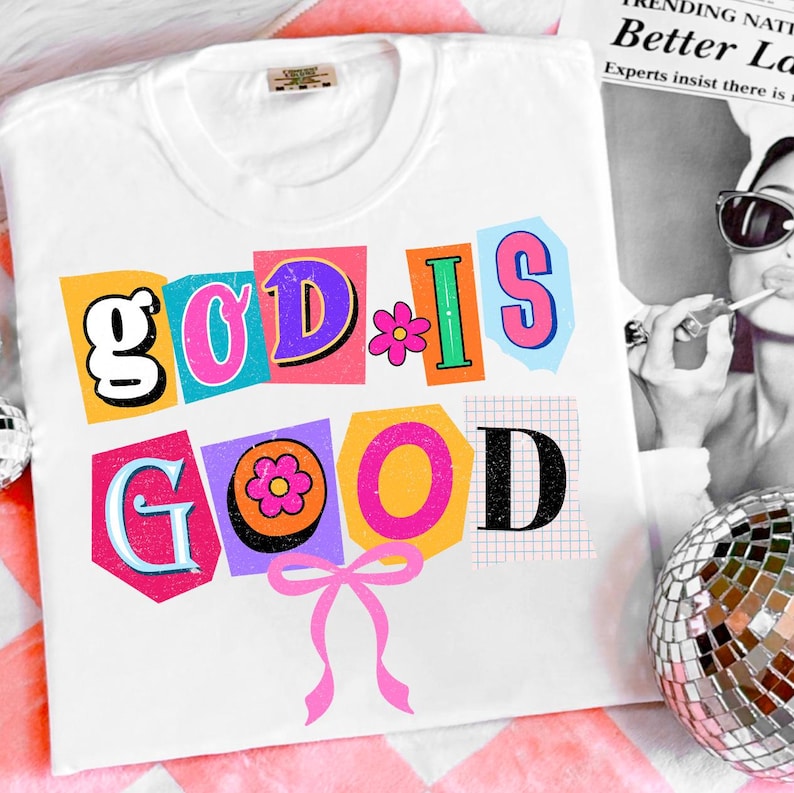 May include: White t-shirt with the phrase "GOD IS GOOD" in colorful, cut-out style letters. The design includes a pink bow and flower accents. The shirt is a casual, everyday wear item.