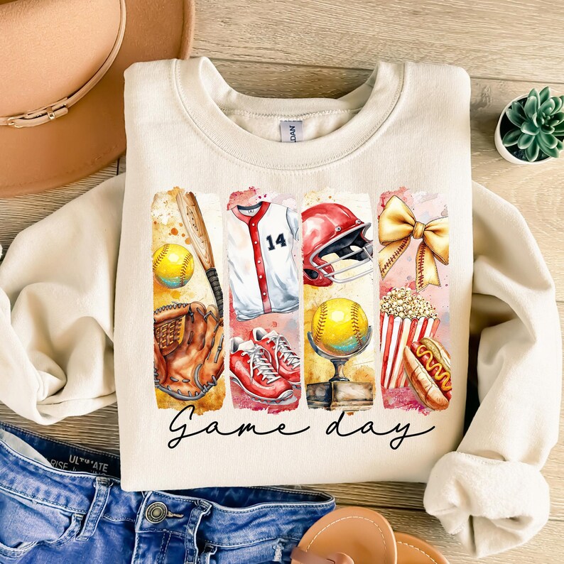 May include: A beige sweatshirt with a watercolor illustration of baseball equipment, including a bat, glove, ball, jersey, helmet, popcorn, and hot dog. The text "Game day" is printed below the illustration.