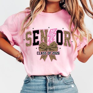 Pu&ograve; includere: T-shirt rosa con la scritta "SENIOR" in nero e stampa leopardata, un fulmine rosa e un fiocco con stampa leopardata. Il testo "CLASS OF 2026" &egrave; sotto il fiocco. La maglietta &egrave; in stile casual.