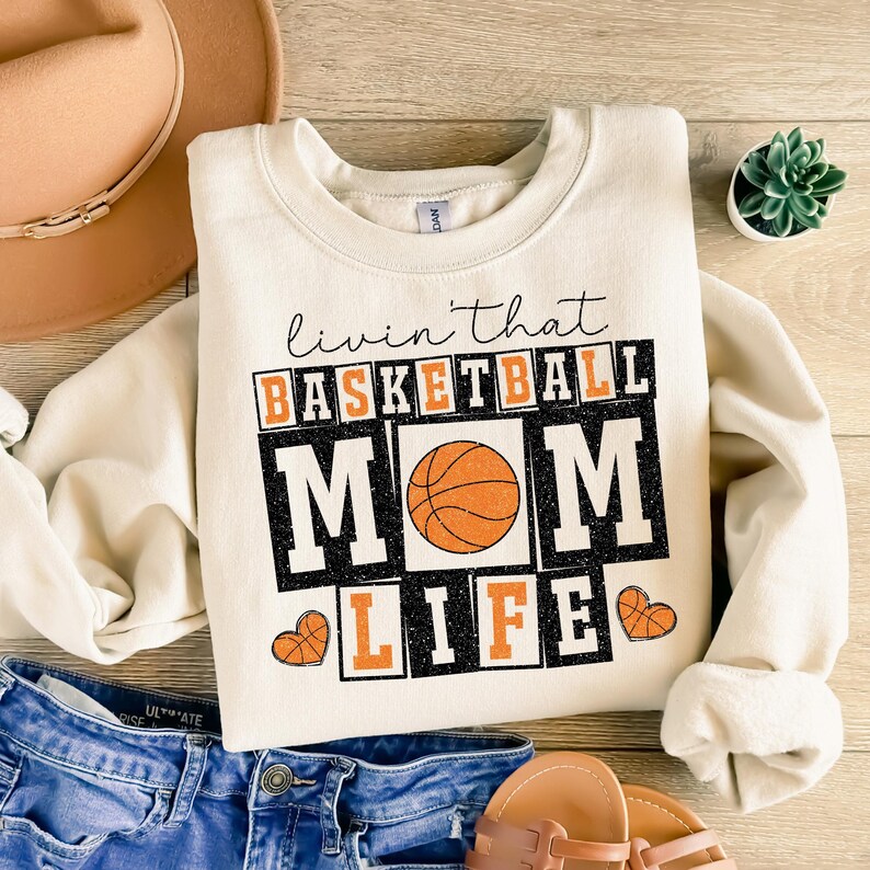 May include: A cream-colored sweatshirt with the text "Livin' that Basketball Mom Life" in black and orange. A basketball graphic is in the center of the text.