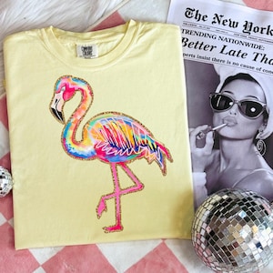 May include: A light yellow t-shirt featuring a colorful flamingo design with pink, blue, yellow, and orange hues. The flamingo has a gold glitter outline. The shirt is next to a newspaper with the headline "TRENDING NATIONWIDE: Better Late Tha".