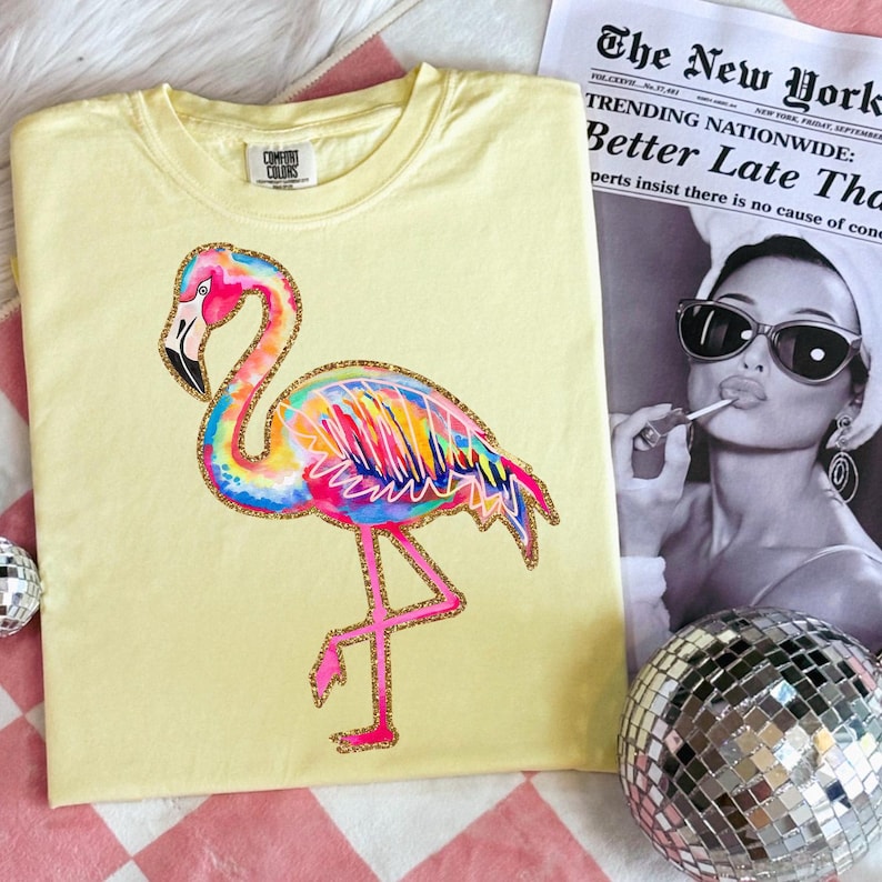 May include: A light yellow t-shirt featuring a colorful flamingo design with pink, blue, yellow, and orange hues. The flamingo has a gold glitter outline. The shirt is next to a newspaper with the headline "TRENDING NATIONWIDE: Better Late Tha".