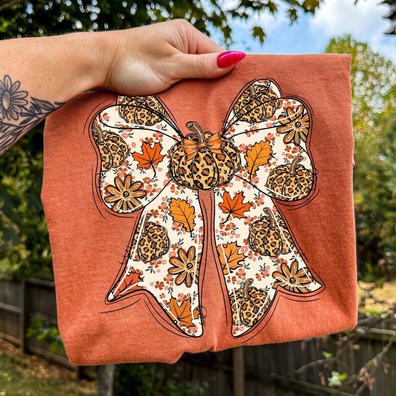 May include: Rust-colored t-shirt featuring a large bow design with a pumpkin and fall leaf pattern. The bow has a leopard print center and is decorated with orange leaves and floral accents, perfect for autumn.