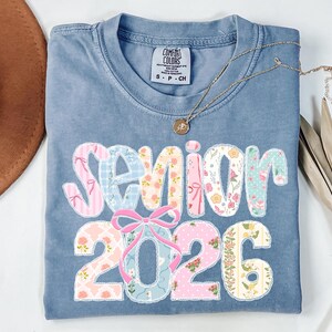 Puede incluir: Una camiseta azul Comfort Colors con la palabra "Senior 2026" en un dise&ntilde;o de patchwork. Las letras y los n&uacute;meros est&aacute;n decorados con telas florales y estampadas. Un collar dorado y un sombrero marr&oacute;n tambi&eacute;n est&aacute;n en la imagen.