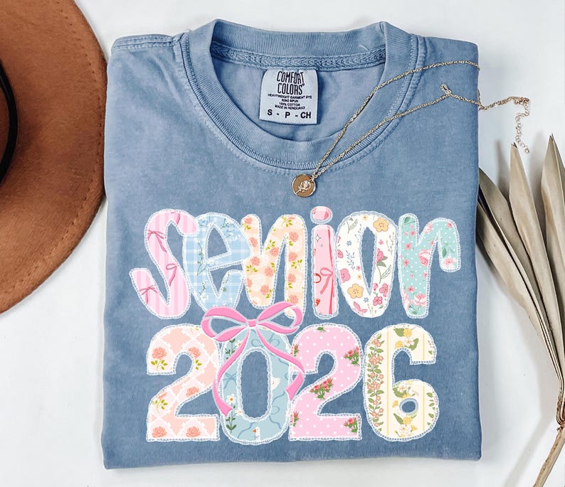 Puede incluir: Una camiseta azul Comfort Colors con la palabra "Senior 2026" en un dise&ntilde;o de patchwork. Las letras y los n&uacute;meros est&aacute;n decorados con telas florales y estampadas. Un collar dorado y un sombrero marr&oacute;n tambi&eacute;n est&aacute;n en la imagen.