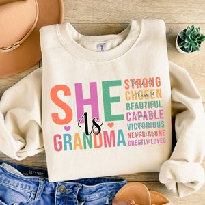 May include: A beige sweatshirt with the text "SHE is GRANDMA" in rainbow colors. The text is surrounded by smaller text in black, including "STRONG, CHOSEN, BEAUTIFUL, CAPABLE, VICTORIOUS, NEVER ALONE, GREATLY LOVED".