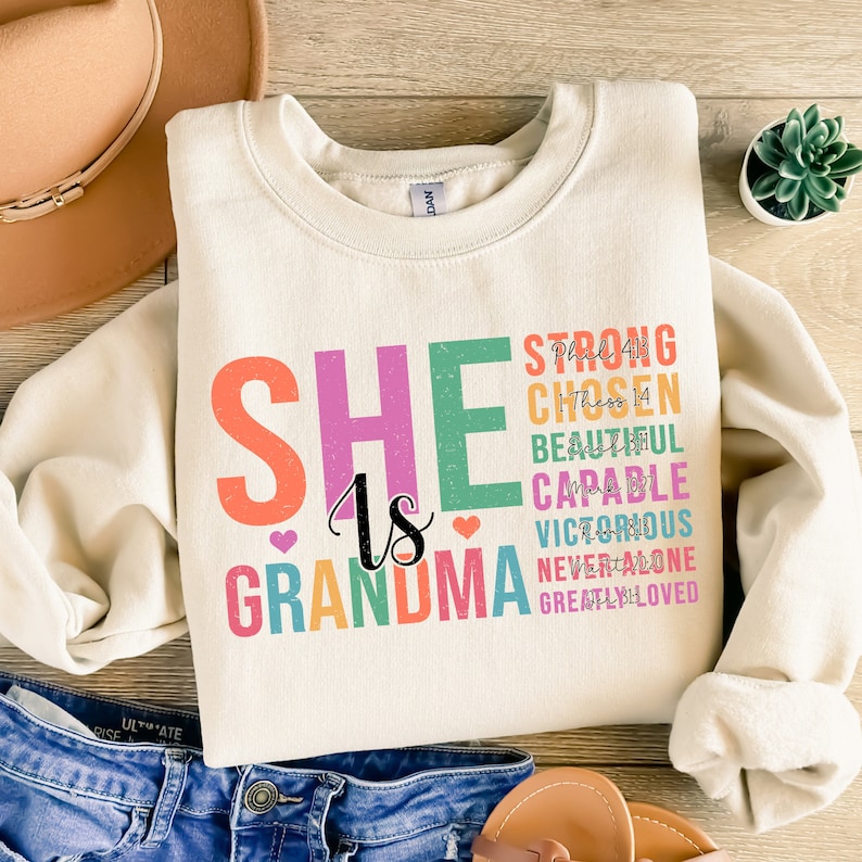 May include: A beige sweatshirt with the text "SHE is GRANDMA" in rainbow colors. The text is surrounded by smaller text in black, including "STRONG, CHOSEN, BEAUTIFUL, CAPABLE, VICTORIOUS, NEVER ALONE, GREATLY LOVED".