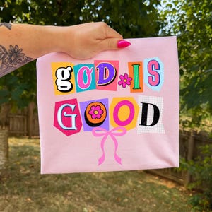 May include: A light pink t-shirt with the words "GOD IS GOOD" in a colorful, collage-style design. The word "GOOD" is below the other words. A pink bow is at the bottom of the design. The shirt is held up by a hand.
