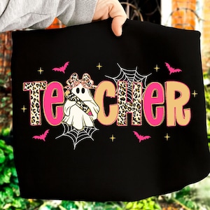 Pu&ograve; includere: T-shirt nero con la scritta "Teacher" in rosa e stampa leopardata beige. Un fantasma con un fiocco leopardato e una matita al centro. Pipistrelli rosa, ragnatele e stelle completano il design.
