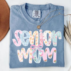 Puede incluir: Una camiseta azul Comfort Colors con las palabras "Senior Mom" en un dise&ntilde;o de patchwork. Las letras presentan patrones florales y a rayas en rosa, azul y blanco. Un collar dorado con un colgante descansa sobre la camiseta.