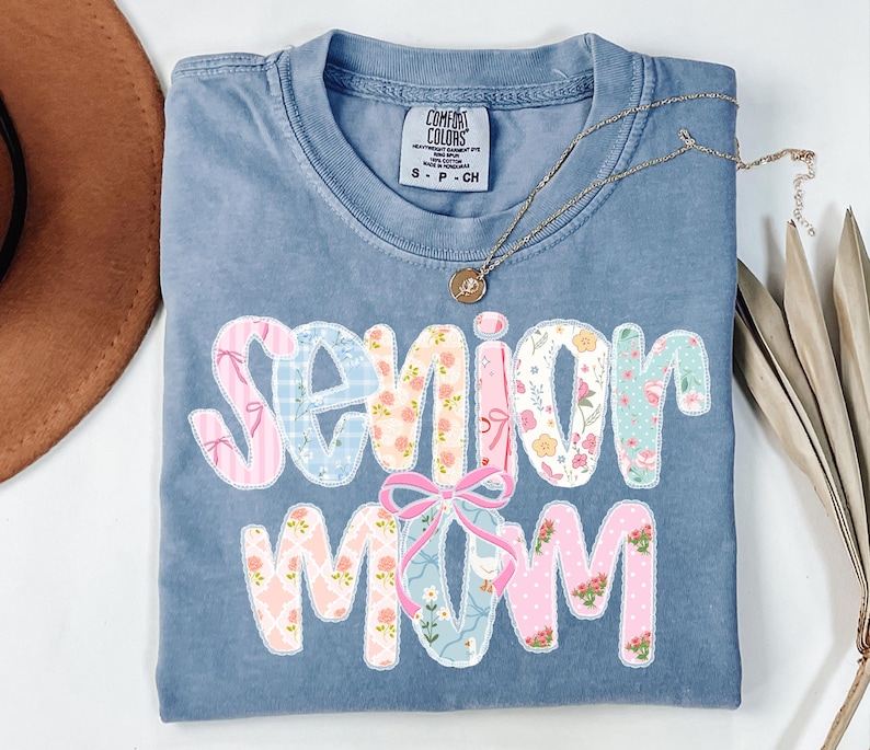 Puede incluir: Una camiseta azul Comfort Colors con las palabras "Senior Mom" en un dise&ntilde;o de patchwork. Las letras presentan patrones florales y a rayas en rosa, azul y blanco. Un collar dorado con un colgante descansa sobre la camiseta.