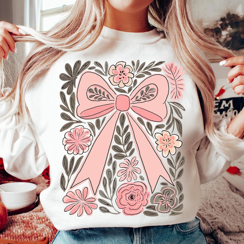 K&ouml;nnte beinhalten: Wei&szlig;es Sweatshirt mit einem gro&szlig;en rosa Schleifendesign, umgeben von rosa und gelben Blumen und grauen Bl&auml;ttern. Die Schleife ist der Mittelpunkt, mit detaillierten floralen und Blattakzenten. Das Design ist auf der Vorderseite des Sweatshirts zentriert.