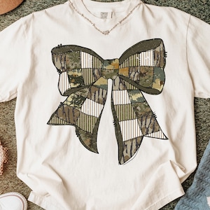 May include: Cream-colored t-shirt featuring a large bow design. The bow is composed of various camouflage patterns and white striped sections, creating a patchwork effect. The shirt has the text "COMFORT COLORS" on the collar.