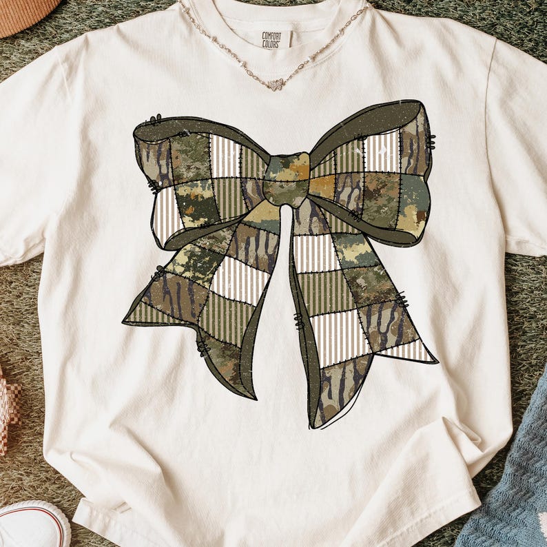 May include: Cream-colored t-shirt featuring a large bow design. The bow is composed of various camouflage patterns and white striped sections, creating a patchwork effect. The shirt has the text "COMFORT COLORS" on the collar.