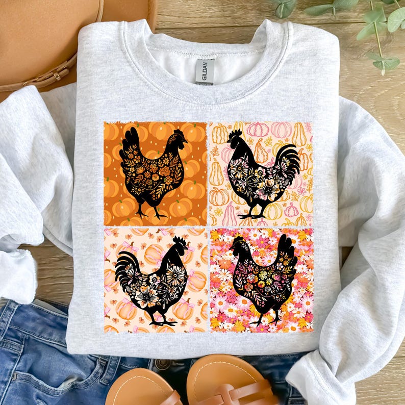 May include: A light gray sweatshirt featuring a four-panel design with black rooster silhouettes filled with floral patterns. The background panels include pumpkins and flowers in orange, pink, and white. The sweatshirt is a Gildan brand.