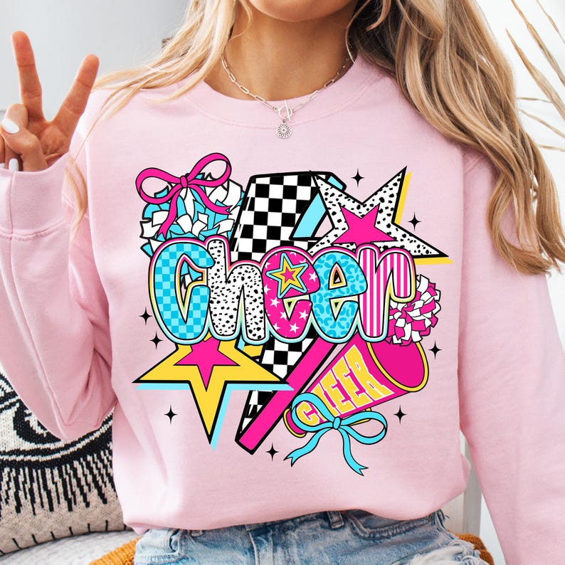 Op de afbeelding: Een lichtroze sweatshirt met een kleurrijke "Cheer"-afbeelding. De afbeelding bevat sterren, een megafoon en een strik. Het woord "Cheer" is in een mix van lettertypen en kleuren, waaronder blauw, roze en zwart.