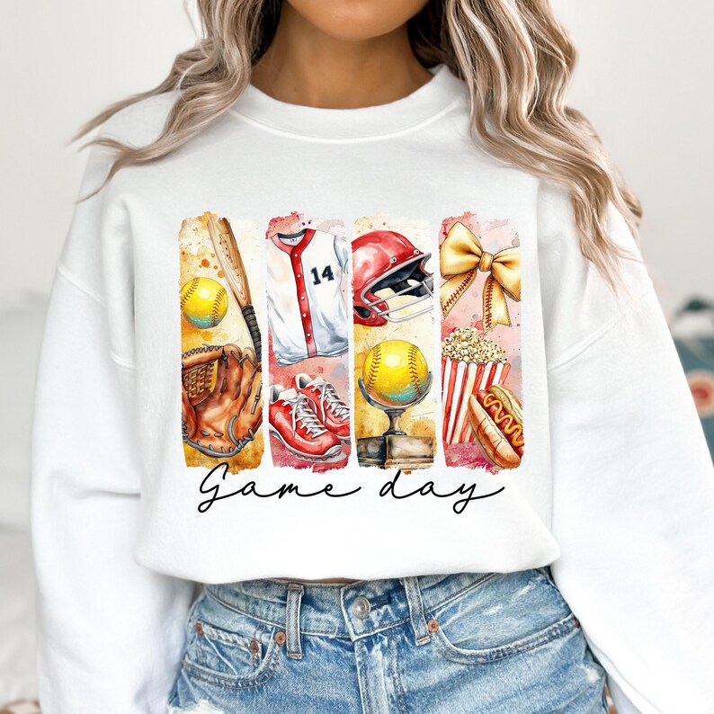 May include: White sweatshirt with a watercolor design of baseball-themed items, including a baseball glove, bat, jersey, helmet, popcorn, hot dog, and a trophy. The text "Game day" is written in cursive below the design.