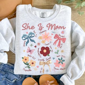 K&ouml;nnte beinhalten: Ein graues Sweatshirt mit dem Text "She Is Mom" in rosa Schrift. Das Sweatshirt ist mit Blumen, Schmetterlingen, Schleifen und Bibelversen verziert.