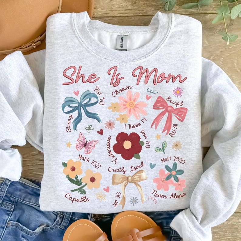 K&ouml;nnte beinhalten: Ein graues Sweatshirt mit dem Text "She Is Mom" in rosa Schrift. Das Sweatshirt ist mit Blumen, Schmetterlingen, Schleifen und Bibelversen verziert.