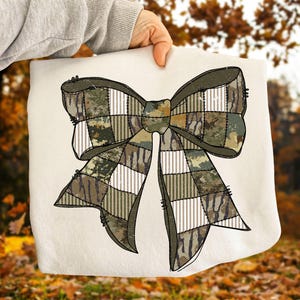 May include: A cream-colored fabric with a large bow design. The bow features a patchwork pattern with camouflage, white, and olive green squares. The bow is outlined in dark brown, and the fabric is held by a person.
