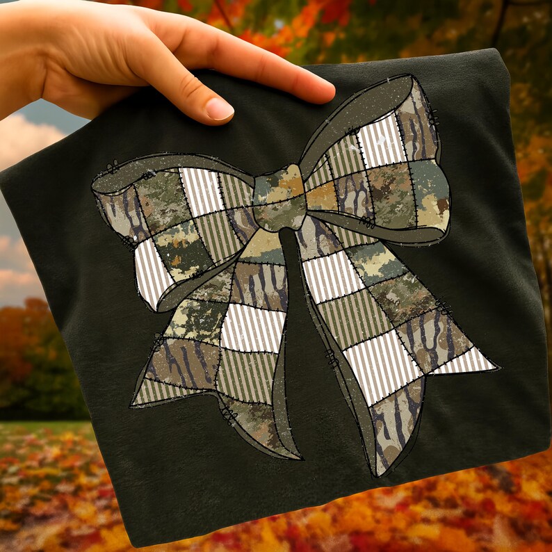 May include: Dark green t-shirt featuring a large bow design. The bow is made of various camouflage patterns, including green, brown, and white stripes and squares. The bow is stitched onto the shirt with a black thread.